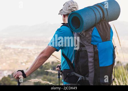 Un randonneur méconnaissable, adulte, homme, caucasien, avec un sac à dos, Tapis, poteaux, vêtus d'un T-shirt bleu, chapeau et pantalon gris est l'observation du paysage Banque D'Images