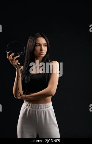 Athlète féminine sérieuse debout avec des écouteurs en studio. Vue portrait d'une jeune fille brunette pensive tenant des écouteurs à la main, tout en regardant loin, isolée sur fond noir. Concept de fitness. Banque D'Images