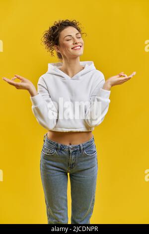 Une jeune fille souriante et détendue essaie de méditer, tout en se tenant à l'intérieur. Vue de face d'une jolie jeune femme en gardant les mains à l'écart, avec des yeux fermés, isolée sur fond jaune de studio. Concept de gestuelle. Banque D'Images