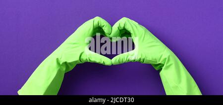 Mains en forme de cœur avec gants en caoutchouc vert sur fond violet. Service de nettoyage de la maison et concept d'entretien ménager. Banque D'Images