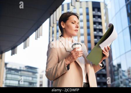 Sérieuse pensive femme d'affaires d'âge moyen debout à l'extérieur et boire du café tout en analysant les données de document dans le presse-papiers Banque D'Images