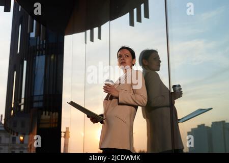 Femme d'affaires, confiante et attentionnée, dans une veste élégante, debout près d'un bâtiment moderne et buvant du café tout en analysant le rapport Banque D'Images