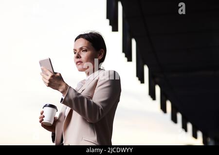 Femme d'affaires sérieuse d'âge moyen dans une veste élégante debout à l'extérieur et utilisant le téléphone tout en obtenant le signal Internet Banque D'Images