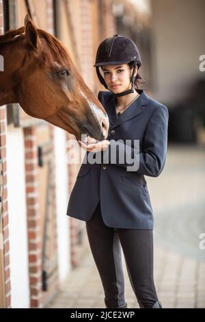 Belle jeune femme jockey se tient vêtu pour la course à côté de son cheval peint rasé. Banque D'Images