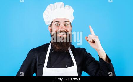 Chef, cuisinier ou boulanger masculin barbu en uniforme, tablier et chapeau de chef pointent le doigt vers le haut. Cuisine, culinaire. Banque D'Images