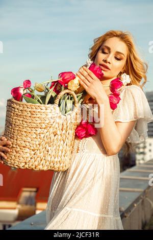 Portrait vertical d'une femme blonde tendre et beutreuse qui prend un panier de fleurs de tulipe qui sent des herbes florales sur le toit Banque D'Images