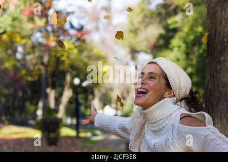 Femme d'âge moyen enthousiaste et aimante, à bras ouverts, regardant les feuilles brunes dans l'air au parc pendant la saison d'automne. La femme mature accueille l'automne Banque D'Images