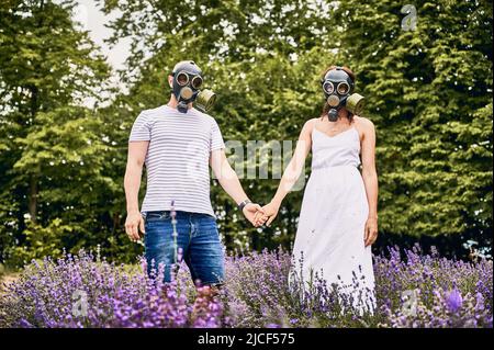 Portrait d'un jeune couple debout dans un champ de lavande portant des masques à gaz, tenant les mains.Personnes souffrant d'allergies et de fleurs de lavande.Concept d'allergie saisonnière Banque D'Images