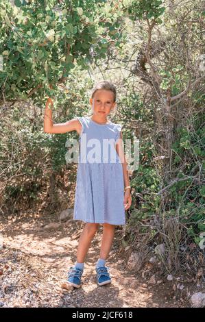 petit garçon fatigué candid fille voyageur et randonneur de huit ans dans une robe à rayures bleues voyage et randonnée d'un chemin de montagne parmi la plante verte de forêt Banque D'Images