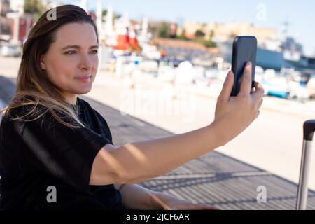 Portrait d'une jeune femme souriante assise près d'un remblai de plage, prenant le selfie dans l'ombre, posant le jour ensoleillé. Banque D'Images