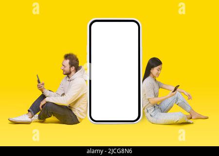 Homme caucasien et fille asiatique assis sur le sol avec des téléphones dans les mains se pencha sur un énorme, géant smartphone blanc écran, publicité d'application mobile isolée sur jaune. Positionnement du produit. Banque D'Images