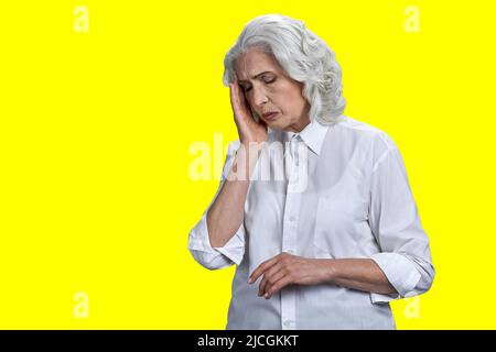 Femme sénior stressée souffrant de migraine sur fond jaune. Concept de maux de tête et de problèmes de santé. Banque D'Images