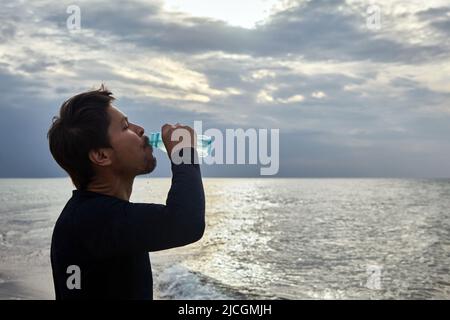 Un homme boit de l'eau après avoir fait du jogging le long de la mer sur fond de ciel de coucher de soleil Banque D'Images