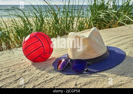 Chapeau de paille, lunettes de soleil et un water-polo rouge sur le sable chaud jaune de la dune sur fond d'herbe verte au bord de la mer. Loisirs actifs c Banque D'Images