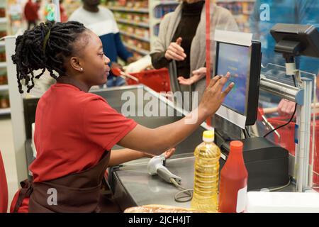 Vue latérale d'une jeune femme noire en uniforme en touchant l'affichage avec les prix des produits alimentaires tout en servant l'acheteur par le comptoir de caisse Banque D'Images