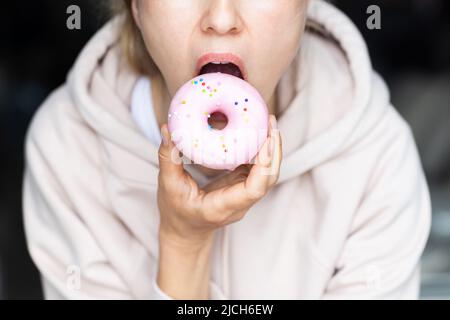 Jeune femme méconnaissable mangeant un beignet rose à la maison dans la cuisine Banque D'Images