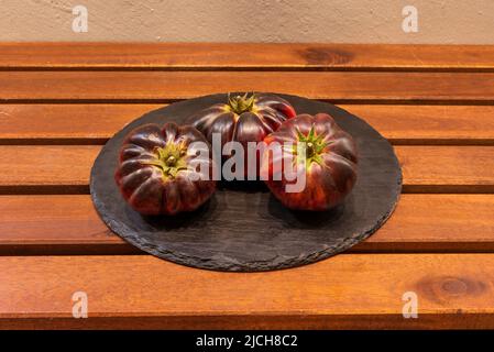 Assiette avec trois tomates de la variété bleue, mûres et prêtes à manger sur une table en bois Banque D'Images