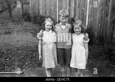 1950s, historique, à l'extérieur dans le jardin arrière, un jeune garçon debout avec ses bras autour de ses deux sœurs plus jeunes, une souriante, avec ses yeux fermés, ..ne regardez pas! Angleterre, Royaume-Uni. Banque D'Images