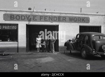 1930s, historique, deux femmes, un petit enfant et un ouvrier de carrosserie de voiture debout pour leur photo à l'entance d'un corps d'auto dans Utah, Mid-West, États-Unis. Un panneau peint à la main sur le bâtiment indique que la carrosserie et l'aile fonctionnent, tandis qu'une automobile de l'époque est garée à l'extérieur sur la piste avec sa roue avant gauche déposée, après avoir fait réparer et peindre son aile - qui recouvre la roue. Les voitures automobiles de cette époque avaient des ailes larges et courbes qui s'étendaient de la porte avant au pare-chocs avant. Banque D'Images