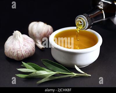 Bouteille versant de l'huile d'olive vierge dans un bol blanc sur fond noir, production d'huile d'olive à l'ail maison Banque D'Images