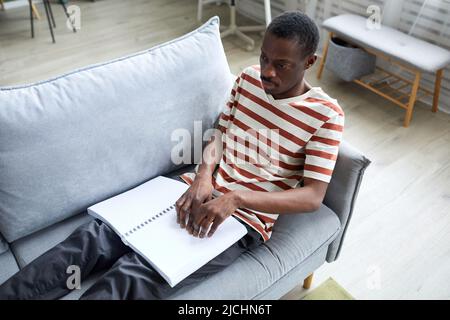 Portrait d'un homme aveugle afro-américain lisant un livre en braille à la maison tout en étant allongé sur un canapé Banque D'Images
