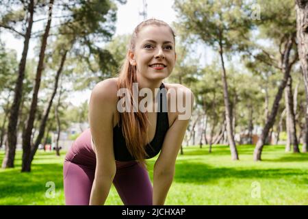 Heureuse femme sportive portant un soutien-gorge de sport debout sur le parc de la ville, debout à l'extérieur et pendu son souffle après une séance de course à pied. Extérieur sp Banque D'Images