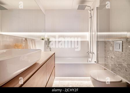 Salle de bains moderne avec articles de sanitaryware en porcelaine blanche, meubles en bois, évier en porcelaine blanche, miroir rectangulaire sans cadre, baignoire avec parcage en verre Banque D'Images