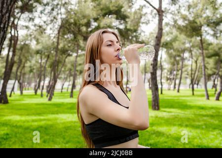 Jeune femme brune portant des vêtements de sport sur le parc de la ville, à l'extérieur de l'eau potable pour se rafraîchir avec le sourire sur le visage. Sports de plein air, vie saine co Banque D'Images