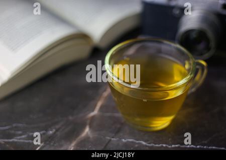 Thé vert infusé dans une tasse transparente près d'un livre ouvert et d'un appareil photo vintage sur fond sombre Banque D'Images