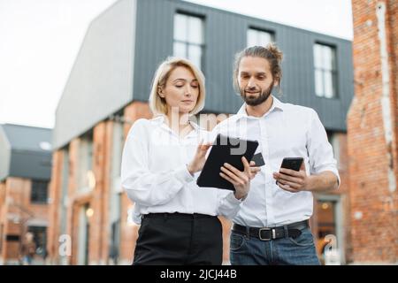 Un couple d'affaires satisfait et attrayant se tenant à l'extérieur près d'un immeuble de bureaux et effectuant un paiement en ligne à l'aide d'une carte de crédit, d'une tablette et d'un smartphone. Vérification du solde du compte, achat de biens concept. Banque D'Images
