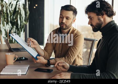 Entrepreneurs de sexe masculin regardant à travers des documents d'affaires, travaillant sur un accord ou un contrat, assis à un bureau dans un bureau moderne Banque D'Images