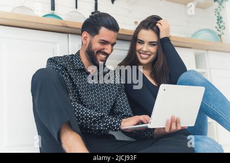 Prise en intérieur dans une cuisine moderne et lumineuse d'un jeune couple gai et interracial, tenant un ordinateur portable et riant. Photo de haute qualité Banque D'Images
