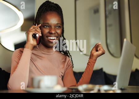 Riant, femme afro-américaine souriante et tortante, blogueur, buvez du café, parlez par téléphone avec vos collègues, travaillez en ligne au café Banque D'Images