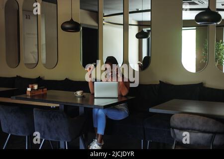Souriante femme d'affaires multiraciale boire un café avec un ordinateur portable et un téléphone dans le café. Dire bonjour aux partenaires. Appels vidéo Banque D'Images