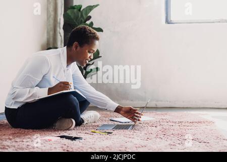 Femme afro-américaine qui étudie avec un ordinateur portable pour se préparer à un examen de test et qui fait ses devoirs au sol à la maison. Prendre des notes. Travaillez à l'intérieur. Calendrier des activités Banque D'Images