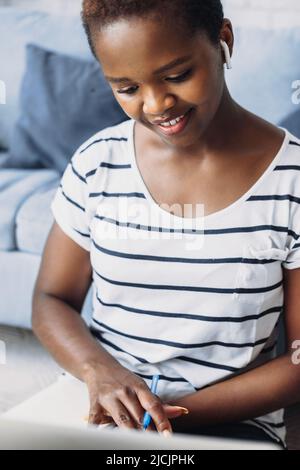 Étudiant afro-américain pensif écrivant des idées dans le journal tout en faisant des devoirs à la maison sur le plancher. Regarder le webinaire d'apprentissage. Rédaction d'une étude de recherche. Banque D'Images