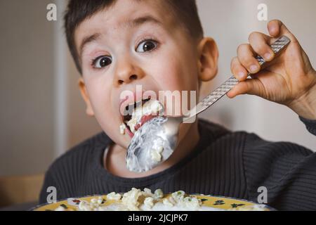 Portrait d'un petit garçon caucasien assis près de la table à la maison mangeant du porridge. Le petit garçon mange de la nourriture saine. Bonheur d'enfance. Alimentation saine et Banque D'Images