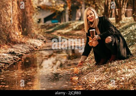 Son élégant croupe blonde près du ruisseau tire sa main à l'eau dans le parc de la ville. Elle est illuminée par la lumière d'automne dorée et tient une tasse avec une boisson chaude dans Banque D'Images