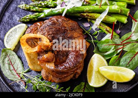 gros plan de l'osso buco braisé, steak de jarret de veau aux asperges grillées, de la lime et des feuilles fraîches du verger suisse sur une assiette noire sur une table en bois sombre Banque D'Images