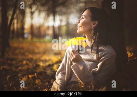 Jeune heureuse femme de course mixte assise près de l'arbre dans le parc d'automne au coucher du soleil Banque D'Images