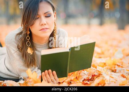 Femme d'asie centrale couchée sur le feuillage d'automne et lisant le livre dans le parc Banque D'Images