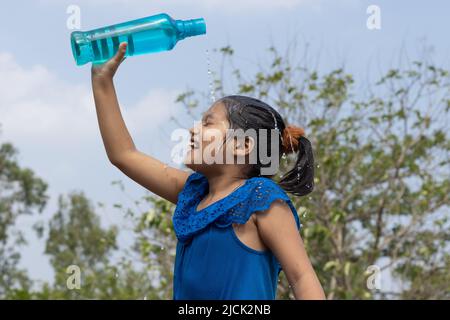 Une fille indienne verse de l'eau sur la tête d'une bouteille en plastique bleu sous le ciel pour se rafraîchir en été Banque D'Images