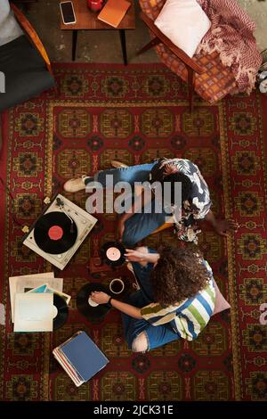 Vue d'ensemble de jeunes couples interculturels en tenues décontractées élégantes assis sur un tapis rouge dans le salon et à l'écoute de disques vynil sur un lecteur de disques Banque D'Images