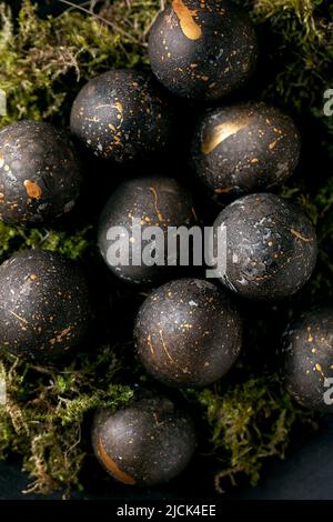 Concept Pâques noir. Œufs noirs de couleur bio avec des taches dorées sur de la mousse sauvage dans un bol en céramique noire sur fond de bois sombre. Vue de dessus, gros plan Banque D'Images