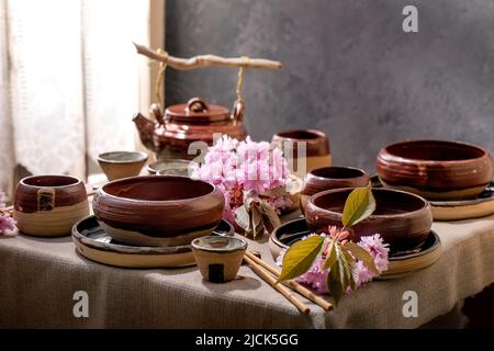 Table de style japonais asiatique avec vaisselle en céramique artisanale vide, bols bruns rugueux, bouilloire et tasses sur la nappe en lin, décorée de sprin rose Banque D'Images
