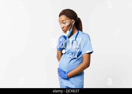 Femme enceinte noire médecin portant un masque facial posant avec un stéthoscope isolé sur fond blanc Banque D'Images