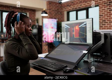Artiste de jeu dans un espace de travail de bureau assis au bureau tout en travaillant sur l'interface de jeu. Niveau employé du département de conception portant des écouteurs sans fil lors de l'analyse de l'environnement numérique Banque D'Images
