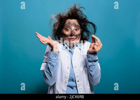 Un biochimiste fou et fou qui regarde un échantillon microscopique après une explosion chimique. Scientifique fou avec des cheveux désordonnés et visage sale regardant confus à la caméra tout en haussant les bras. Banque D'Images