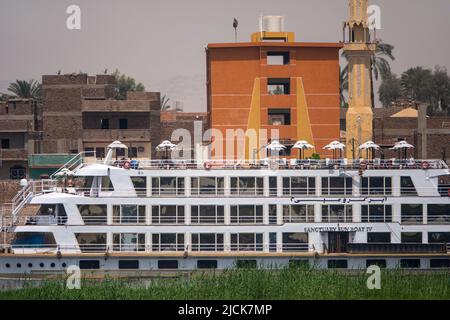 Bateau de croisière sur le Nil à bord d'un appareil photo derrière une île avec bande verte d'herbe avec mur de bâtiments derrière comme toile de fond Banque D'Images