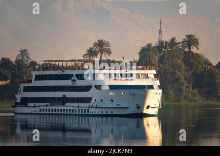 Bateau de croisière sur le Nil naviguant sur l'eau calme avec des reflets de palmiers, mosquée, montagne et navire dans la lumière du matin Banque D'Images